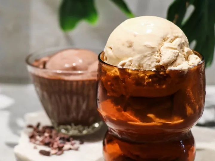 Scoop of gelato in a glass with cacao nibs and biscuit crumbs beside a cup of chocolate gelato