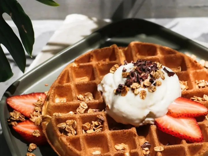 Freshly made waffle topped with gelato, granola and sliced strawberries on a green plate